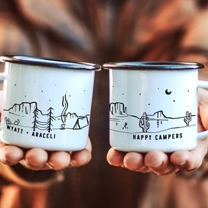 Könnte beinhalten: Zwei weiße Emailletassen mit schwarzen Griffen und schwarzen Illustrationen. Eine Tasse trägt die Aufschrift "WYATT + ARACELI" und zeigt eine Wüstenlandschaft mit einem Lagerfeuer, einem Zelt und Bäumen. Die andere Tasse trägt die Aufschrift "HAPPY CAMPERS" und zeigt eine Wüstenlandschaft mit einem Kaktus und einer Mondsichel.