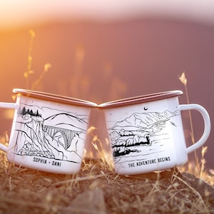 K&ouml;nnte beinhalten: Zwei wei&szlig;e Emailletassen mit schwarzen Illustrationen. Eine Tasse zeigt eine Bergszene mit einer Br&uuml;cke und dem Text "Sophia + Dani". Die andere Tasse zeigt eine Bergszene mit einem Fluss und dem Text "The Adventure Begins".