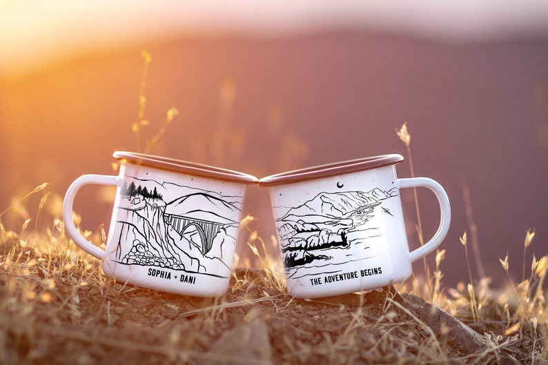 K&ouml;nnte beinhalten: Zwei wei&szlig;e Emailletassen mit schwarzen Illustrationen. Eine Tasse zeigt eine Bergszene mit einer Br&uuml;cke und dem Text "Sophia + Dani". Die andere Tasse zeigt eine Bergszene mit einem Fluss und dem Text "The Adventure Begins".