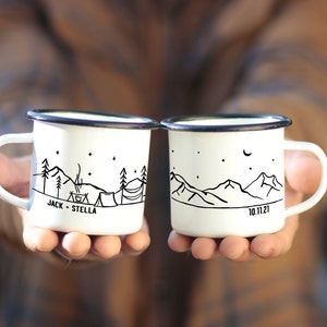 Könnte beinhalten: Zwei weiße Emailletassen mit schwarzen Rändern und Griffen. Eine Tasse zeigt eine Bergszene mit einem Lagerfeuer, einem Zelt und dem Text "JACK + STELLA". Die andere Tasse zeigt eine Bergszene mit einem Halbmond und dem Text "10.11.21".