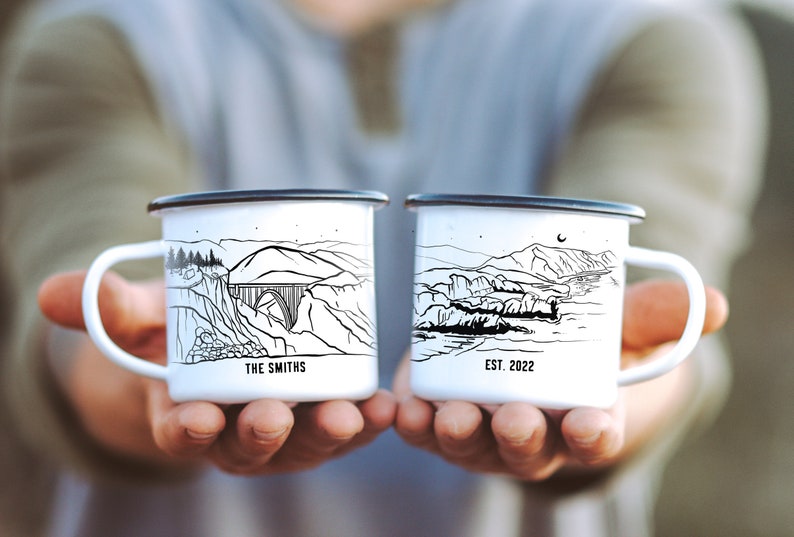 K&ouml;nnte beinhalten: Zwei wei&szlig;e Emailletassen mit schwarzen Umrissen. Die Tasse links hat eine Zeichnung einer Br&uuml;cke und den Text "THE SMITHS". Die Tasse rechts hat eine Zeichnung einer K&uuml;ste und den Text "EST. 2022".