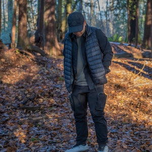 May include: A person wearing a black puffer vest, a black long-sleeved shirt, black cargo pants, and a black baseball cap with a white circle logo. The person is standing in a forest with fallen leaves on the ground.