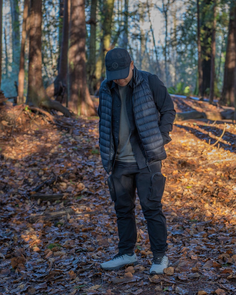 May include: A person wearing a black puffer vest, a black long-sleeved shirt, black cargo pants, and a black baseball cap with a white circle logo. The person is standing in a forest with fallen leaves on the ground.
