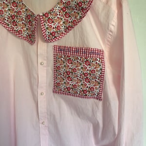 Peut inclure: Une chemise boutonnée rose avec un grand col à motif floral blanc, rouge et floral. La chemise a une poche plaquée avec une bordure en vichy rouge et blanc et un motif floral.