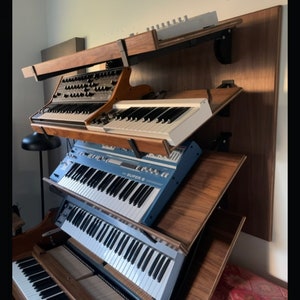 May include: A wall-mounted wooden shelf unit with four levels, each holding a different keyboard instrument. The top level has a white keyboard, the second level has a blue keyboard with the text "SUPER 6" on it, the third level has a brown keyboard, and the bottom level has a black and white piano keyboard.