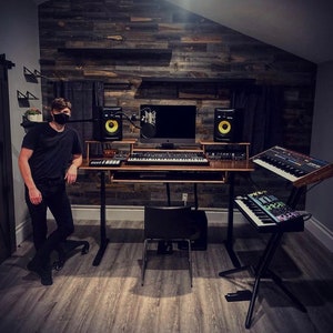 May include: A home music studio with a person standing in front of a wooden wall with a desk and multiple keyboards. The desk has a computer monitor, two speakers, and a microphone. The person is wearing a black shirt and a black face mask.