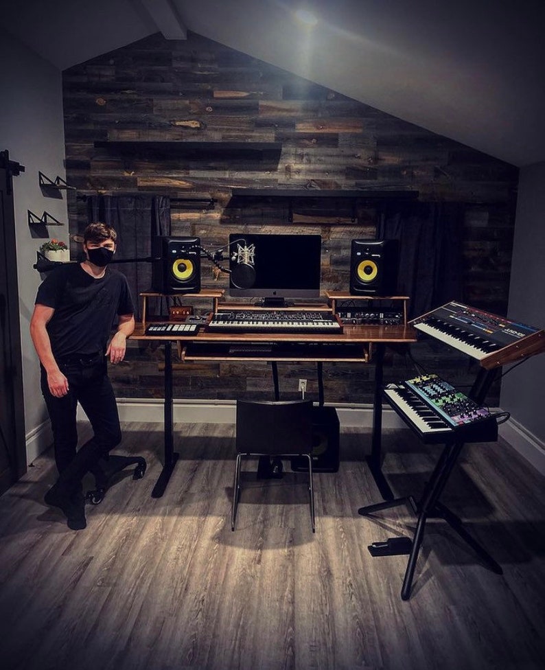 May include: A home music studio with a person standing in front of a wooden wall with a desk and multiple keyboards. The desk has a computer monitor, two speakers, and a microphone. The person is wearing a black shirt and a black face mask.