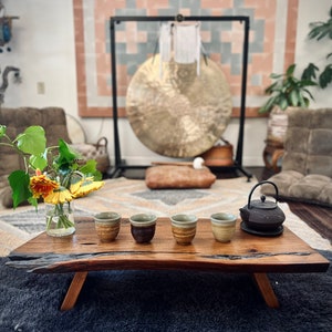 May include: A wooden table with five small ceramic cups and a black cast iron teapot on a wooden tray. The table is set on a gray rug in front of a large gong hanging from a stand.
