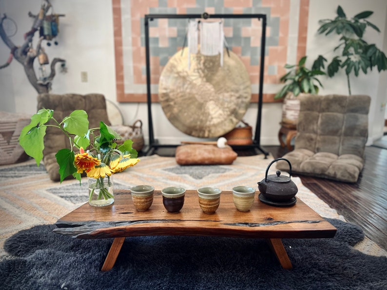May include: A wooden table with five small ceramic cups and a black cast iron teapot on a wooden tray. The table is set on a gray rug in front of a large gong hanging from a stand.