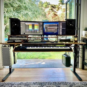 May include: A black computer desk with a keyboard, two monitors, and a pair of black speakers. The desk is set up in a home office with a view of a backyard through a large window.