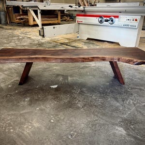 May include: A long, dark brown live edge wood table with two angled legs. The table is made of natural wood with a unique grain pattern. The table is sitting on a concrete floor.