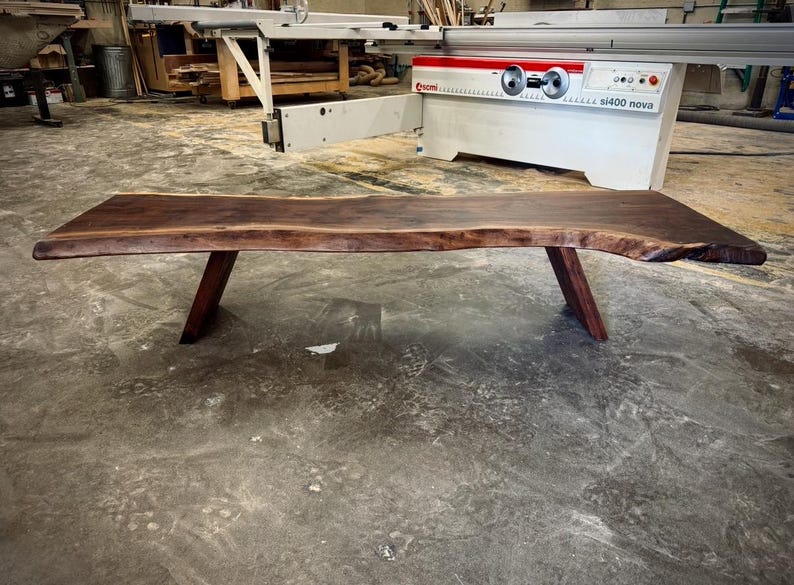 May include: A long, dark brown live edge wood table with two angled legs. The table is made of natural wood with a unique grain pattern. The table is sitting on a concrete floor.
