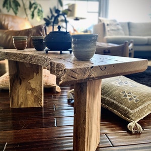 May include: A rustic wooden table with a natural wood finish and a live edge. The table has two legs and is sitting on a hardwood floor. There are three ceramic teacups and a black teapot on the table.