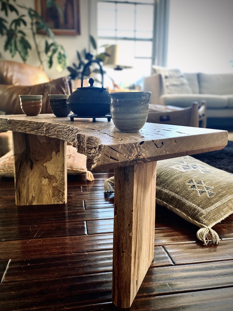 May include: A rustic wooden table with a natural wood finish and a live edge. The table has two legs and is sitting on a hardwood floor. There are three ceramic teacups and a black teapot on the table.