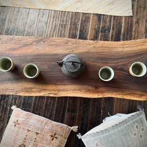 May include: A wooden table with a live edge, featuring four small ceramic cups and a black cast iron teapot. The table is set on a wooden floor with a rug in the background.