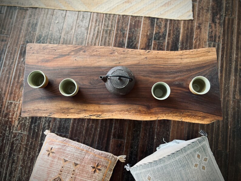 May include: A wooden table with a live edge, featuring four small ceramic cups and a black cast iron teapot. The table is set on a wooden floor with a rug in the background.