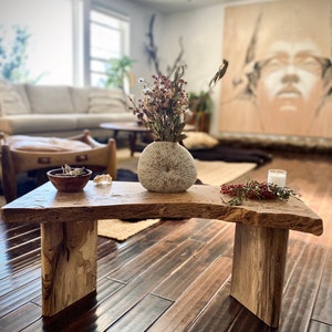 May include: A rustic wooden bench with a natural wood finish. The bench has two legs and a curved top. A white ceramic vase with dried flowers sits on the bench, along with a small wooden bowl and a white candle.