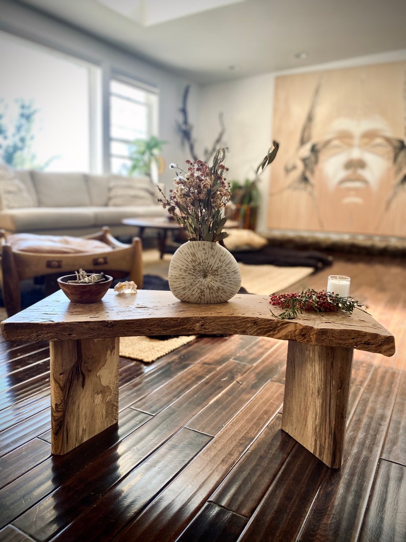 May include: A rustic wooden bench with a natural wood finish. The bench has two legs and a curved top. A white ceramic vase with dried flowers sits on the bench, along with a small wooden bowl and a white candle.