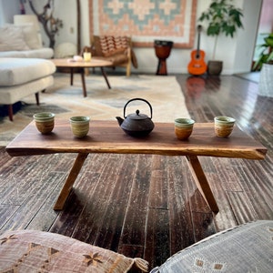 May include: A rustic wooden coffee table with a live edge top and two legs. Four small ceramic teacups and a black cast iron teapot are arranged on the table. The table is in a living room with a rug, a couch, and a guitar.