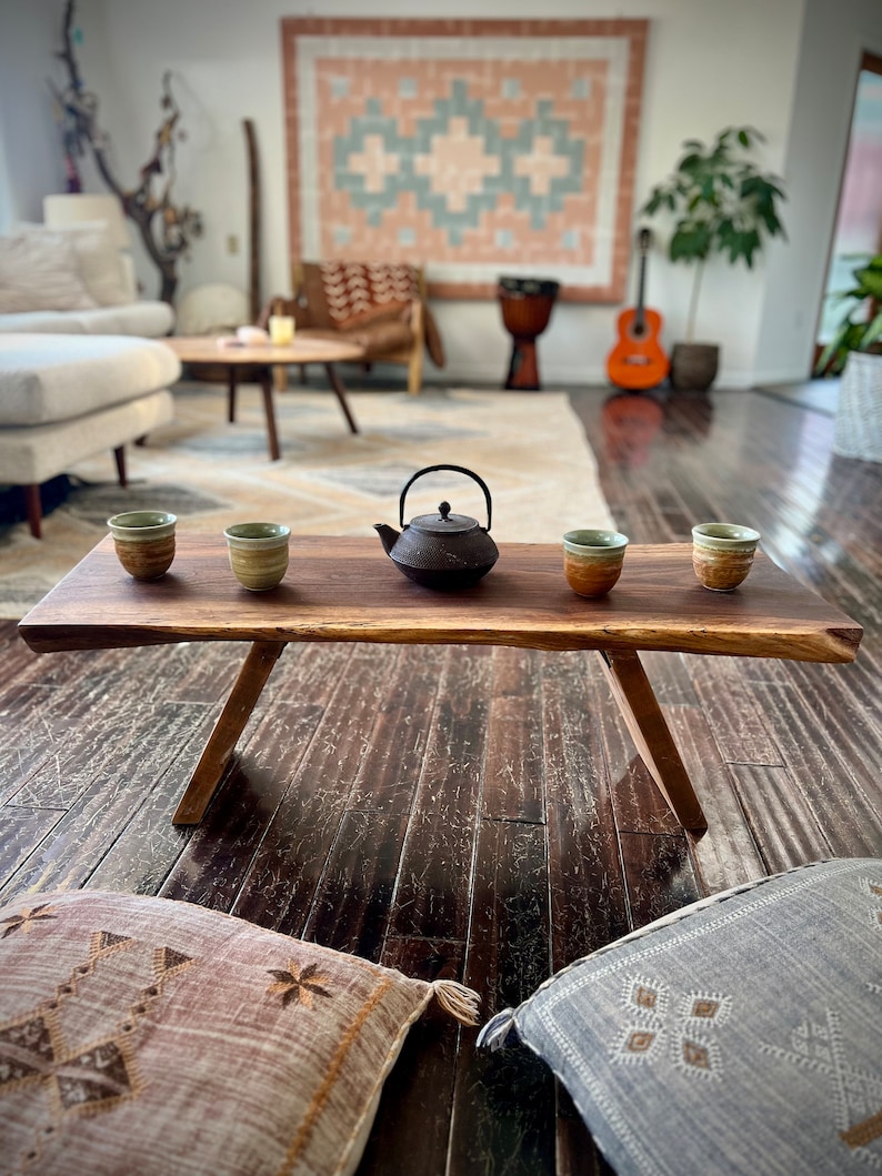 May include: A rustic wooden coffee table with a live edge top and two legs. Four small ceramic teacups and a black cast iron teapot are arranged on the table. The table is in a living room with a rug, a couch, and a guitar.