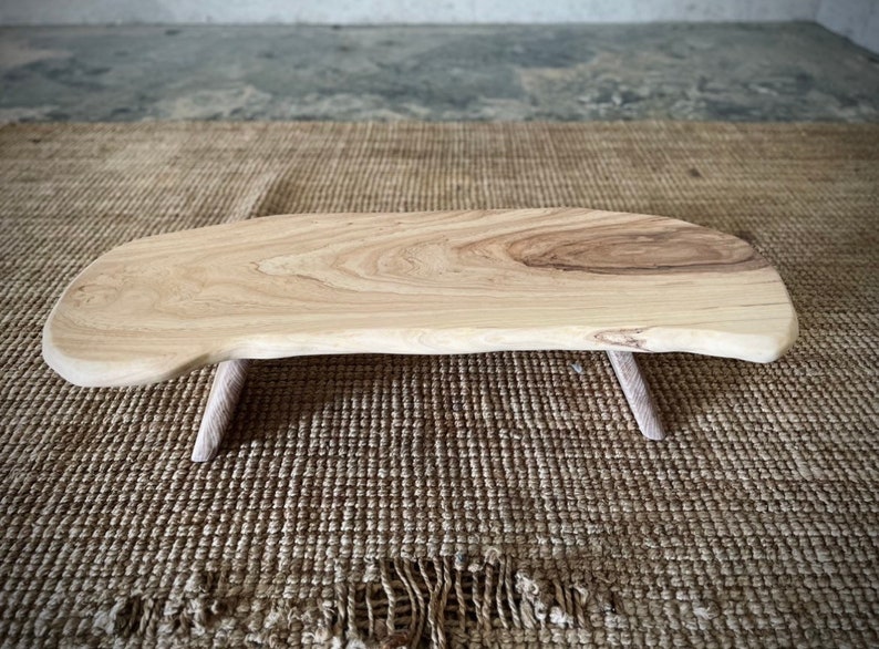 May include: A natural wood serving tray with a live edge and two small wooden legs. The tray is light brown in color and has a smooth, polished finish.