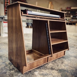May include: A wooden keyboard stand with a built-in shelf and drawer. The stand is made of dark wood and has a sleek, modern design.