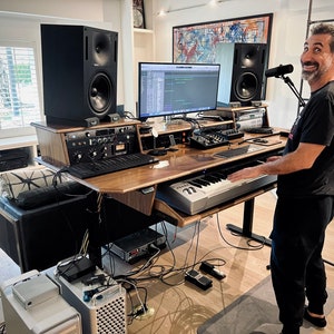 May include: A man stands in a home recording studio, playing a keyboard. The studio features a large wooden desk with a computer monitor, a microphone, and two black speakers. The desk is surrounded by various electronic equipment and cables.