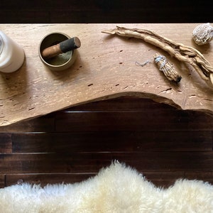 May include: A wooden table top with a candle, a singing bowl, a piece of driftwood, a bundle of sage, and a crystal. The table top is light brown with a natural wood grain.