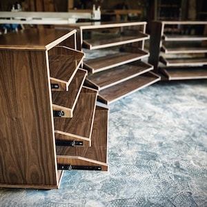 May include: A corner shelf unit made of wood with a dark walnut finish. The shelves are angled and have a pull-out drawer. The unit is made of multiple pieces that can be assembled to create a custom configuration.