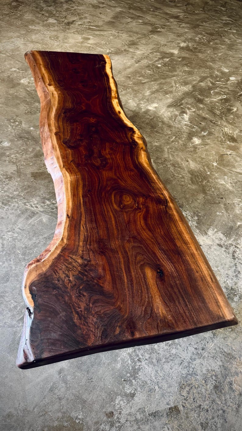 May include: A large, live edge slab of wood with a rich, dark brown color and a natural, wavy grain pattern. The wood has a smooth, polished finish.