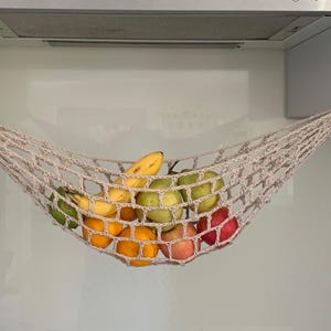 May include: A beige, crocheted fruit hammock suspended from hooks, filled with a variety of fresh fruits. Oranges, apples, bananas, and green pears are visible. The hammock is hanging in a kitchen setting.
