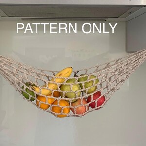 Può includere: Un'amaca per frutta in macramè beige piena di vari frutti, tra cui arance, mele, pere e banane. L'amaca è sospesa a ganci attaccati a un mobile da cucina. Il testo "PATTERN ONLY" è visualizzato sopra l'amaca.
