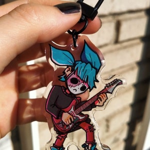 Puede incluir: Llavero acrílico transparente con un personaje de dibujos animados con pelo azul, una máscara y una camisa negra tocando una guitarra roja. El llavero tiene un cierre negro y se sostiene contra un fondo de ladrillos.