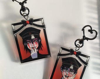 May include: Two acrylic keychains with heart-shaped clasps. Each keychain features a framed illustration of a person in a black hat and uniform, with red accents and a bow. The background is red.