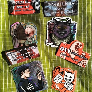 Puede incluir: Una colección de varios paquetes de pegatinas sobre una superficie verde a cuadros. Los paquetes presentan diseños con texto como "SILENT HILL", "FEAR & HUNGER", "CRY OF FEAR" y "OFF". Las pegatinas muestran ilustraciones de personajes y temas.