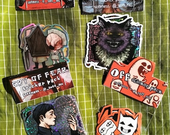 May include: A collection of various sticker packs on a green checkered surface. The packs feature designs with text like "SILENT HILL", "FEAR & HUNGER", "CRY OF FEAR", and "OFF". The stickers showcase illustrations of characters and themes.