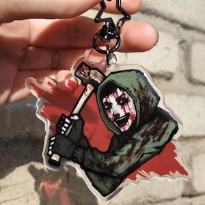 Könnte beinhalten: Ein klarer Acryl-Schlüsselanhänger mit einer Figur in einer grünen Kapuzenjacke, die einen Hammer vor einem rot blutbespritzten Hintergrund hält. Der Schlüsselanhänger hat einen schwarzen sternförmigen Verschluss.