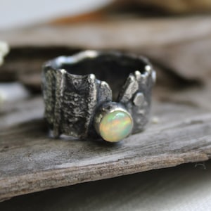 May include: A silver ring with a textured band and a white opal gemstone. The ring is sitting on a piece of driftwood.