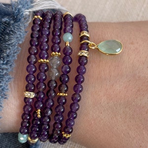 May include: A set of five amethyst gemstone bracelets with gold accents and a single green gemstone pendant. The bracelets are stacked on a person's wrist.