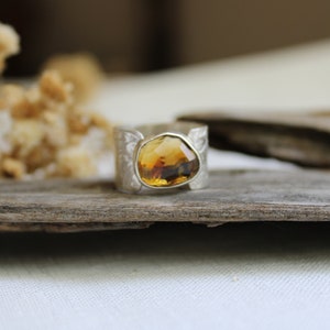 May include: A silver ring with a large, faceted, amber-colored gemstone set in the center. The ring is on a piece of weathered wood.