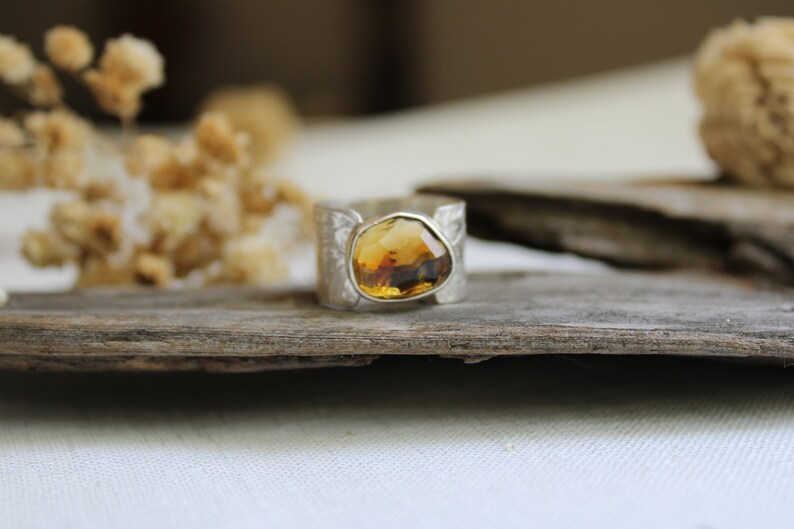 May include: A silver ring with a large, faceted, amber-colored gemstone set in the center. The ring is on a piece of weathered wood.
