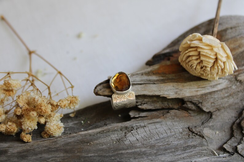 May include: A silver ring with an amber gemstone set in a textured band. The ring is displayed on a piece of driftwood with dried flowers.