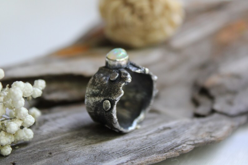 May include: A silver ring with a textured, organic design. The ring features a white opal gemstone set in the center.