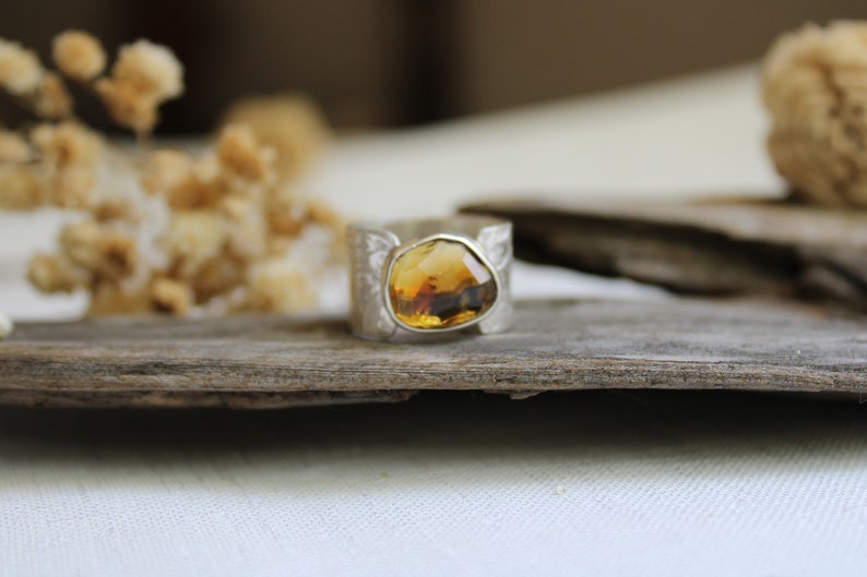 May include: A silver ring with a large, faceted, amber-colored gemstone set in the center. The ring is on a piece of weathered wood.