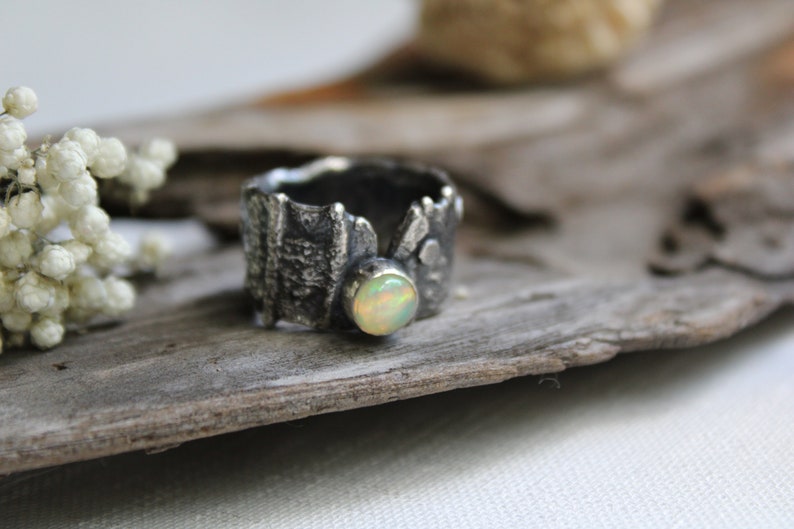 May include: A silver ring with a textured band and a white opal gemstone. The ring is sitting on a piece of driftwood.