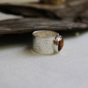 May include: A silver ring with an ornate design and a brown gemstone. The ring is set on a white fabric background.