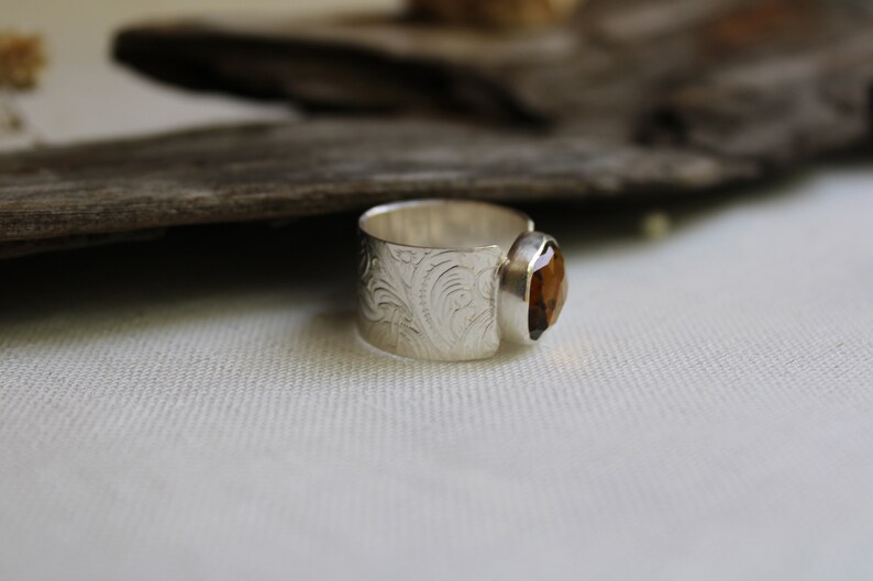 May include: A silver ring with an ornate design and a brown gemstone. The ring is set on a white fabric background.