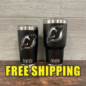 May include: Two black Yeti tumblers with a silver New Jersey Devils logo engraved on each. The tumblers are on a wooden surface with the text "FREE SHIPPING" in yellow letters.