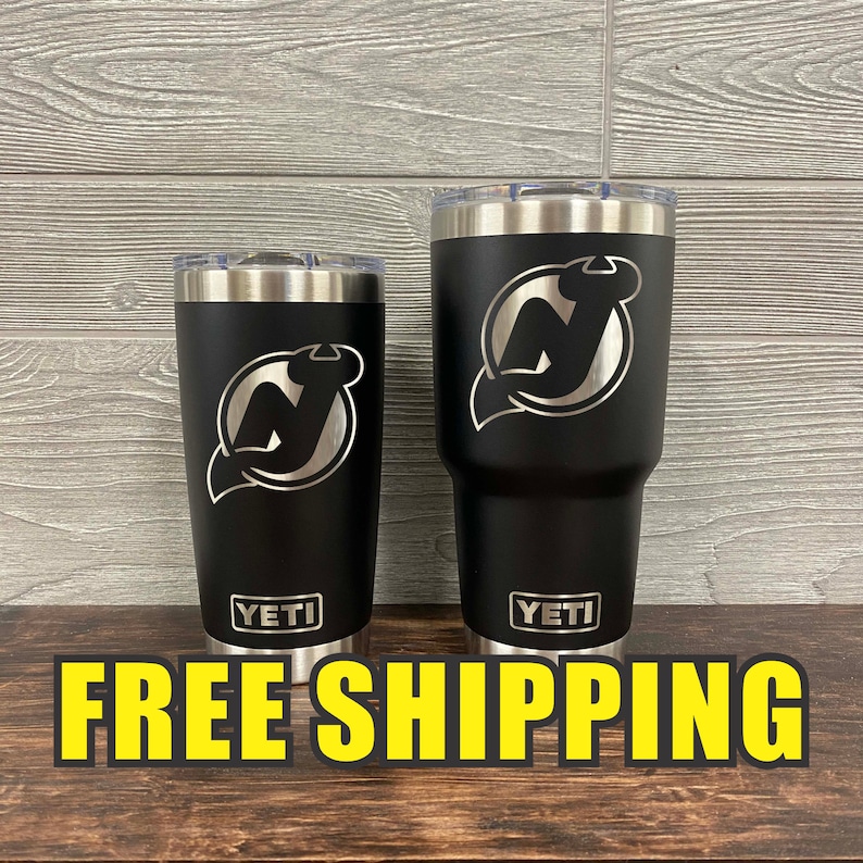 Puede incluir: Dos vasos Yeti negros con un logotipo plateado de los New Jersey Devils grabado en cada uno. Los vasos est&aacute;n sobre una superficie de madera con el texto "ENV&Iacute;O GRATIS" en letras amarillas.