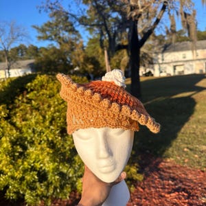 May include: A crocheted hat shaped like a pie, featuring a brown crust and a filling of orange and white yarn. The hat is displayed on a white mannequin head against a backdrop of green foliage and a blue sky.